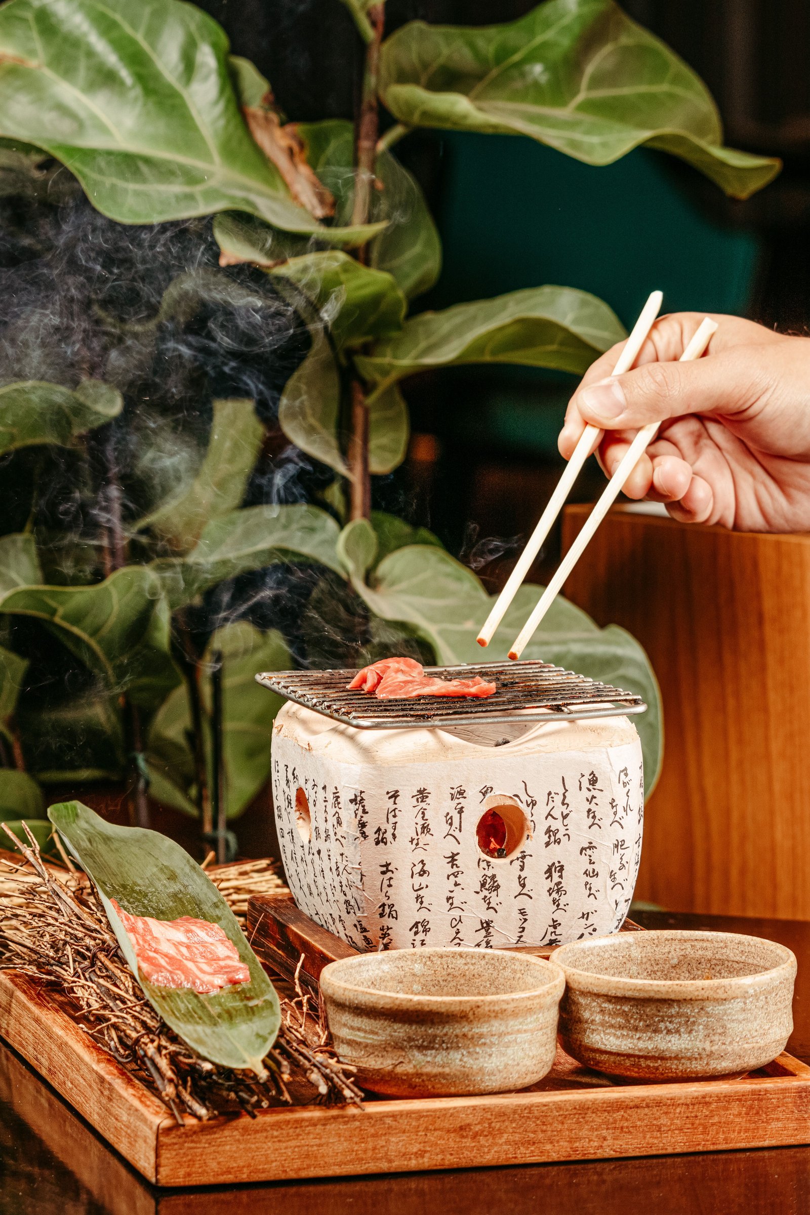 Hands turning beef on a ceramic tabletop grill beside chopsticks
